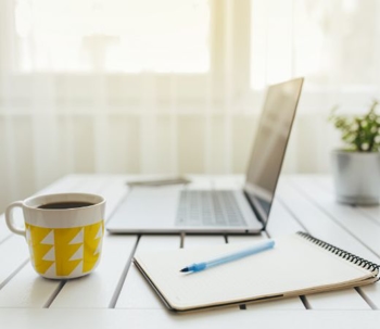 Laptop, cup of coffee, and notepad on top of a desk.
